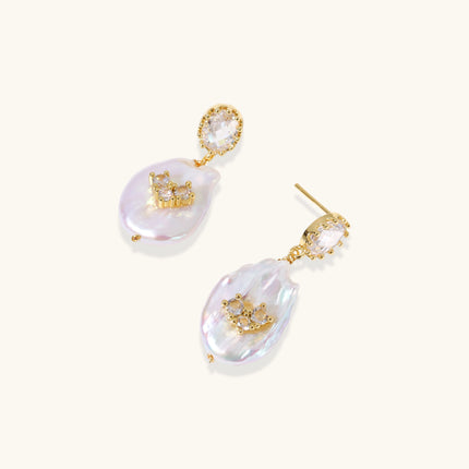 Gold-plated drop earrings with iridescent baroque pearls and crystal flower accents on cream background