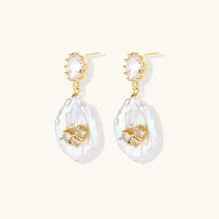Gold-plated drop earrings with iridescent baroque pearls and crystal flower accents on cream background