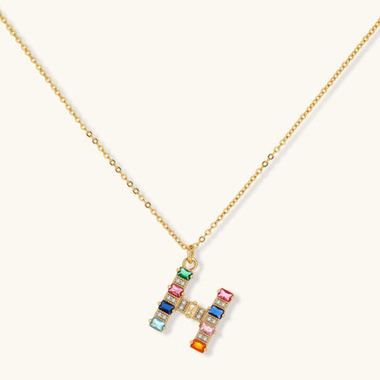 Gold-plated H initial pendant necklace and stud earrings set with multicolor baguette zirconia stones on cream background.