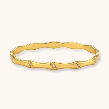 Gold-toned wavy bangle with bamboo-style segments and CZ crystal clusters on cream background. 18K plated stainless steel.